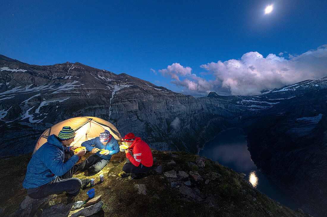 Three men hikers enjoying dinner outside a tent above lake Limmernsee at dusk, Canton of Glarus, Switzerland, Europe
