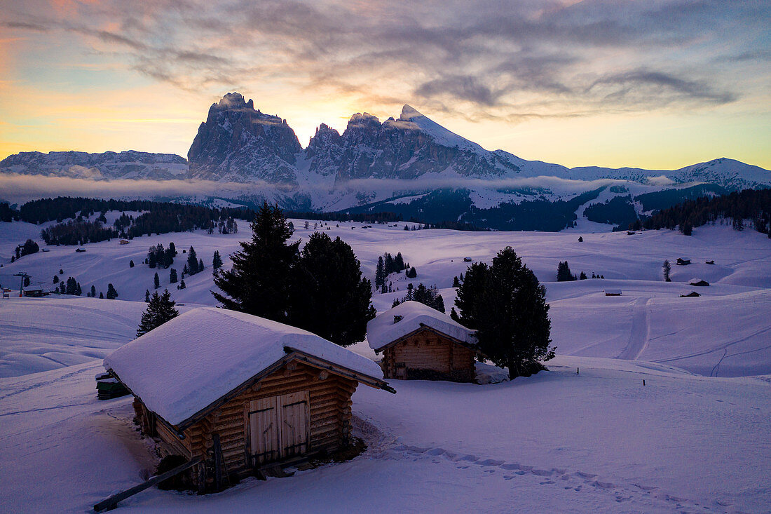 Holzhütten bedeckt mit Schnee mit Sassopiatto und Sassolungo im Hintergrund im Morgengrauen, Seiser Alm, Dolomiten, Südtirol, Italien, Europa