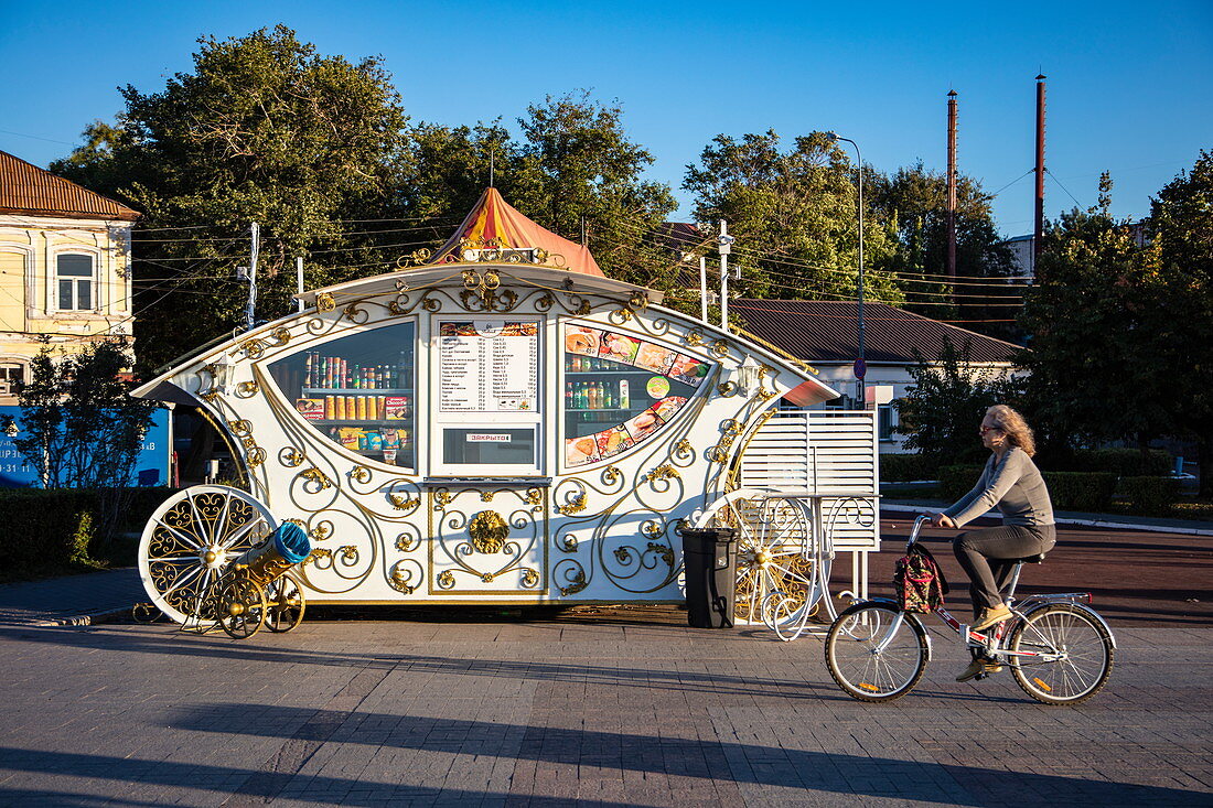 Woman on bicycle in front of snack bar in the design of a carriage along promenade on the bank of Volga River, Astrakhan, Astrakhan District, Russia, Europe