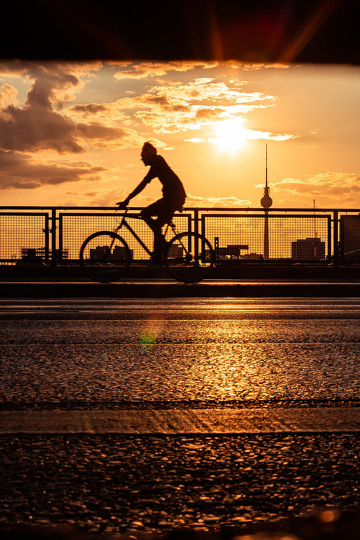 Schattenbild des Mannes, der auf Brücke mit Fernsehturm Berlin im Hintergrund während des Sonnenuntergangs radelt