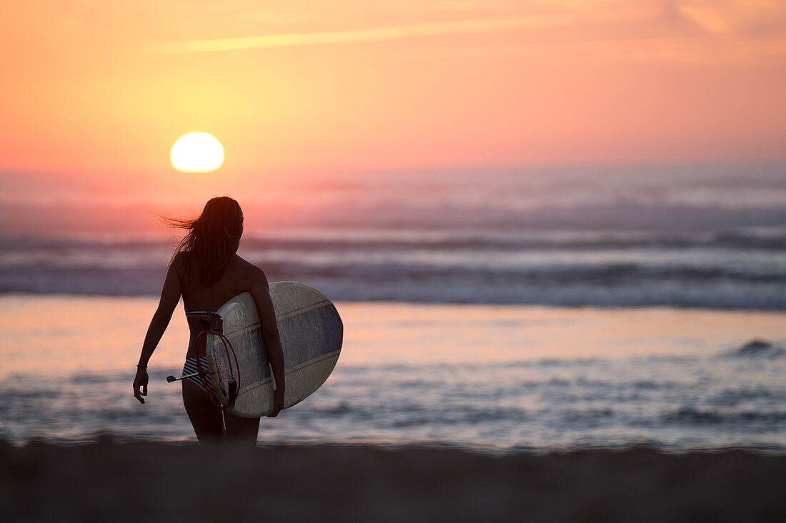 Surferin geht mit Surfbrett am Strand im Sonnenuntergang,  Portugal