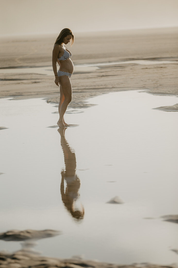 schwangere Frau steht im Abendlicht am Strand