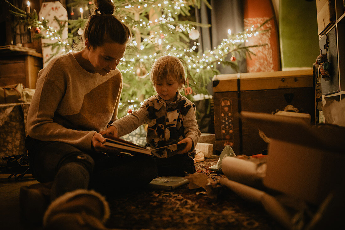 Kleiner Junge schaut mit seiner Mutter Buch vorm Weihnachtsbaum an