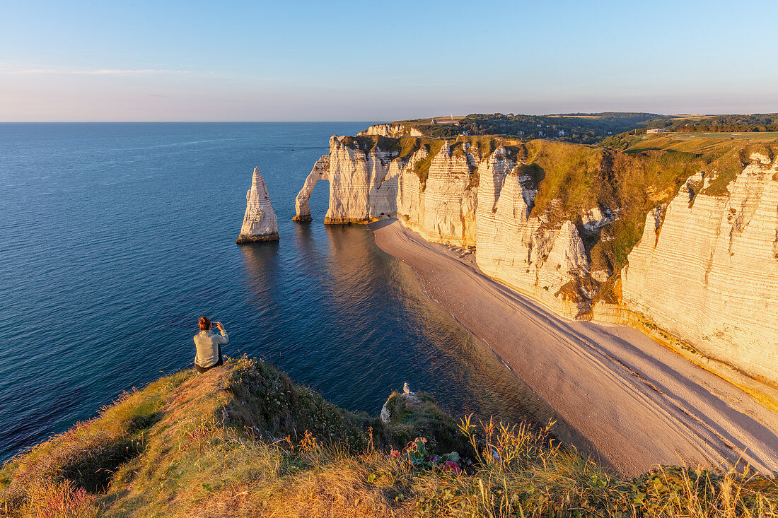 A woman sits on the Alabaster Coast and looks at the Porte d'Aval rock arch and the Aiuille rock needle near Étretat, Normandy, France.