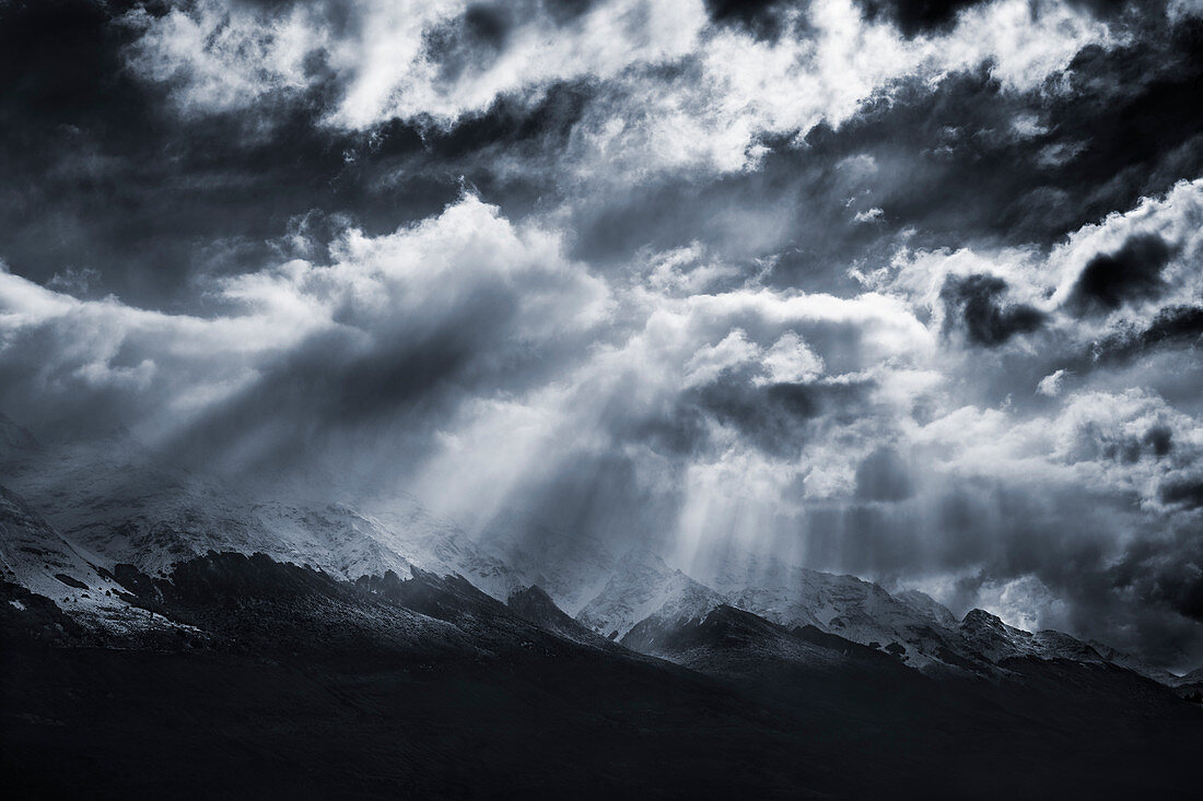 Rays of sunlight filter through dramatic clouds onto a snow topped mountain range outside of Queenstown, South Island, New Zealand.