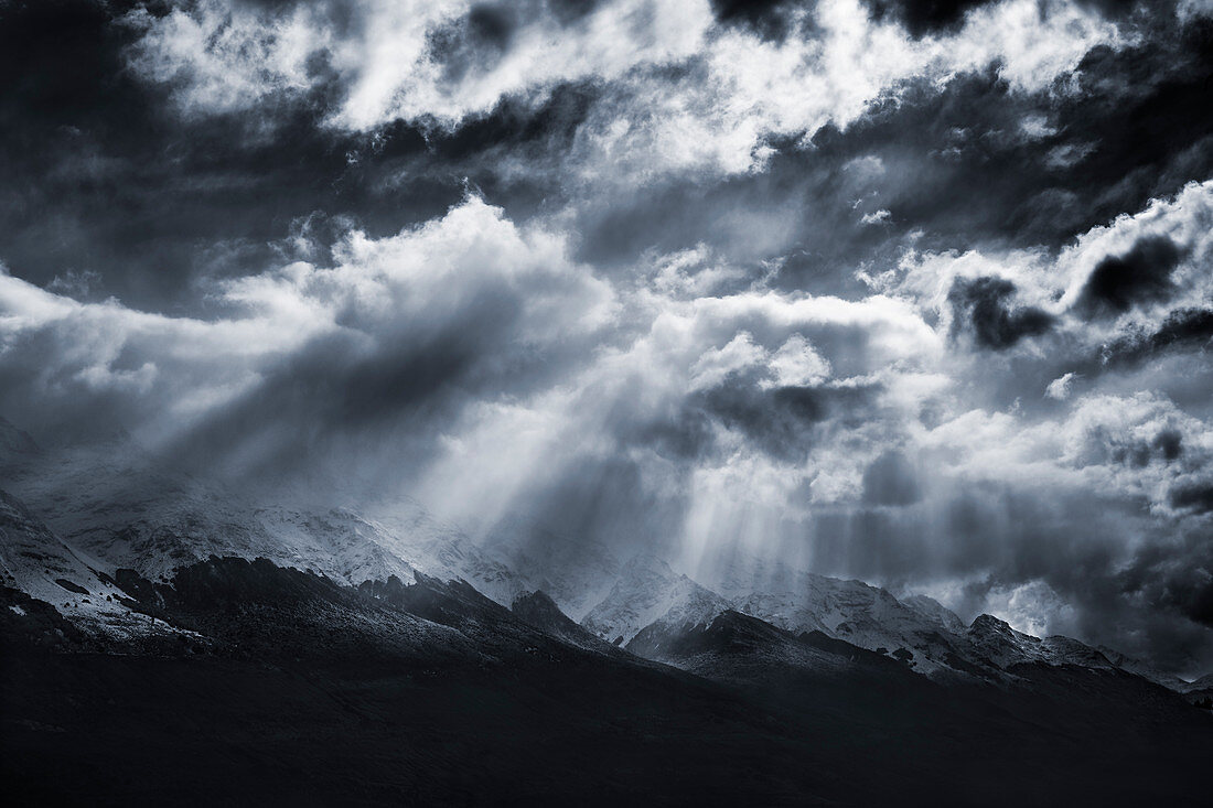 Sonnenstrahlen fallen durch dramatische Wolken auf eine schneebedeckte Bergkette außerhalb von Queenstown, Südinsel, Neuseeland