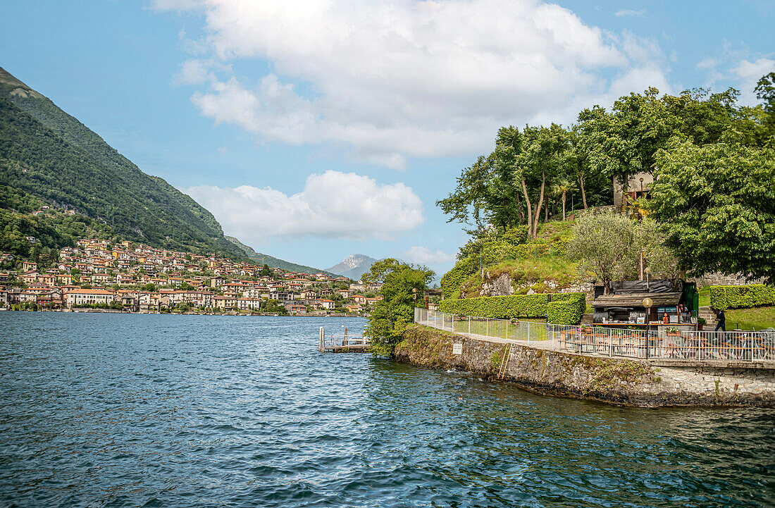 Schiffsanlegestelle an der Seepromenade von Isola Comacina am Comer See von der Seeseite gesehen, mit der Stadt Ossuccio im Hintergrund, Lombardei, Italien 