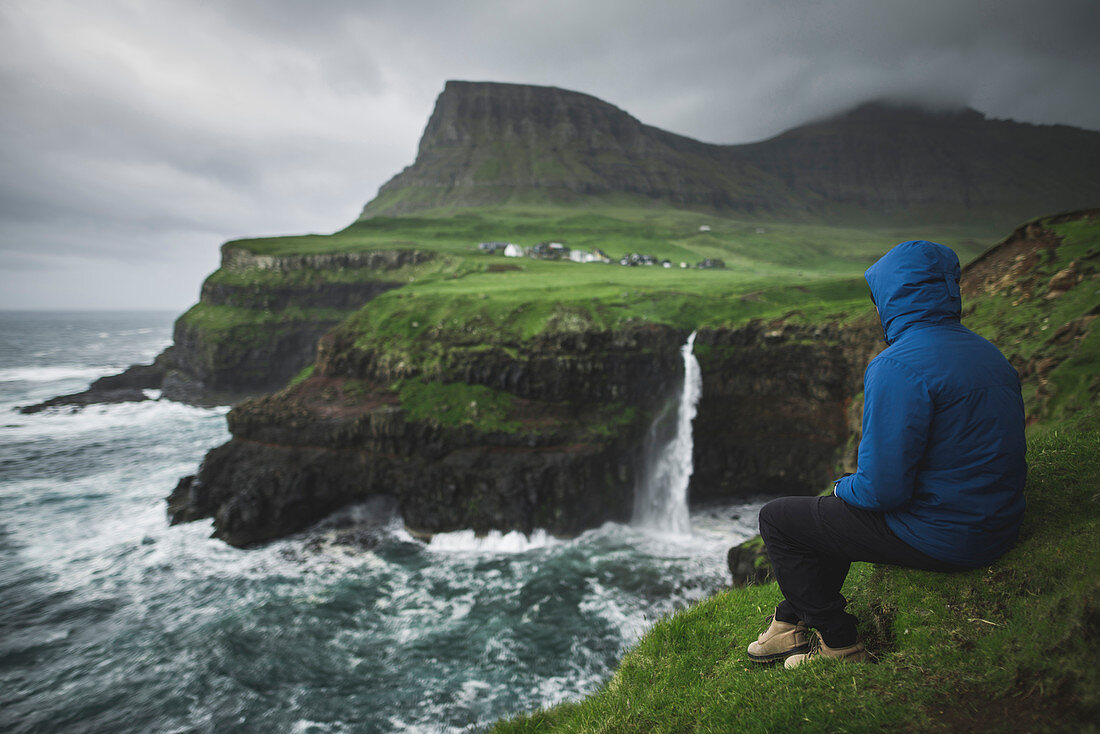 Dänemark, Färöer, Dorf Gasadalur, M? Lafossur Wasserfall, Mann sitzt am Rand der Klippe und Blick auf Mulafossur Wasserfall