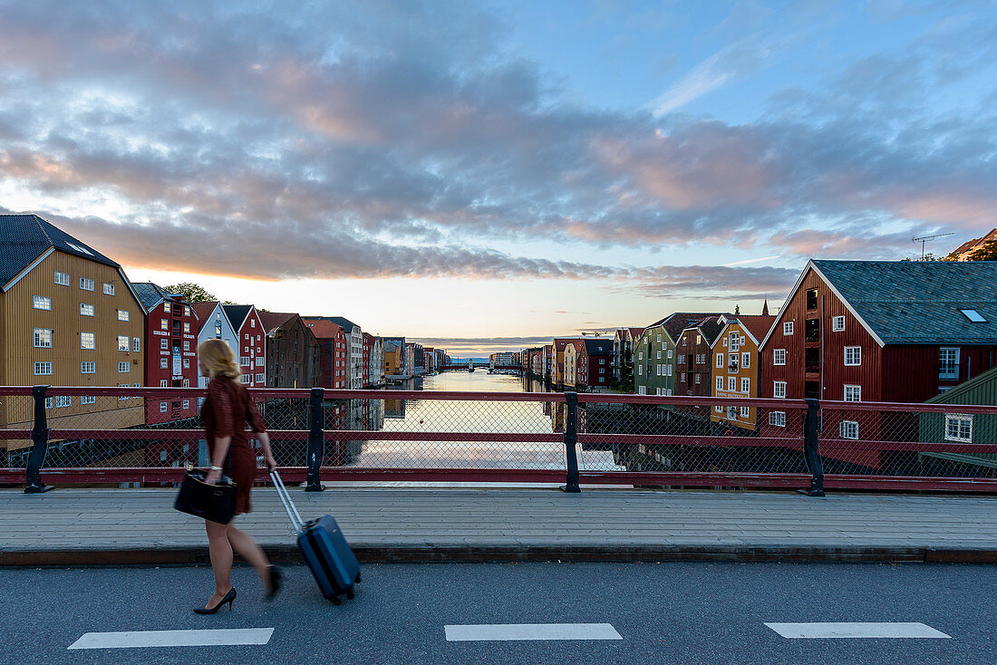 Frau mit Rollkoffer auf der Brücke Bybrua am Fluss Nidelv, Alte Lagerhäuser entlang der Nidelva, Trondheim, Norwegen