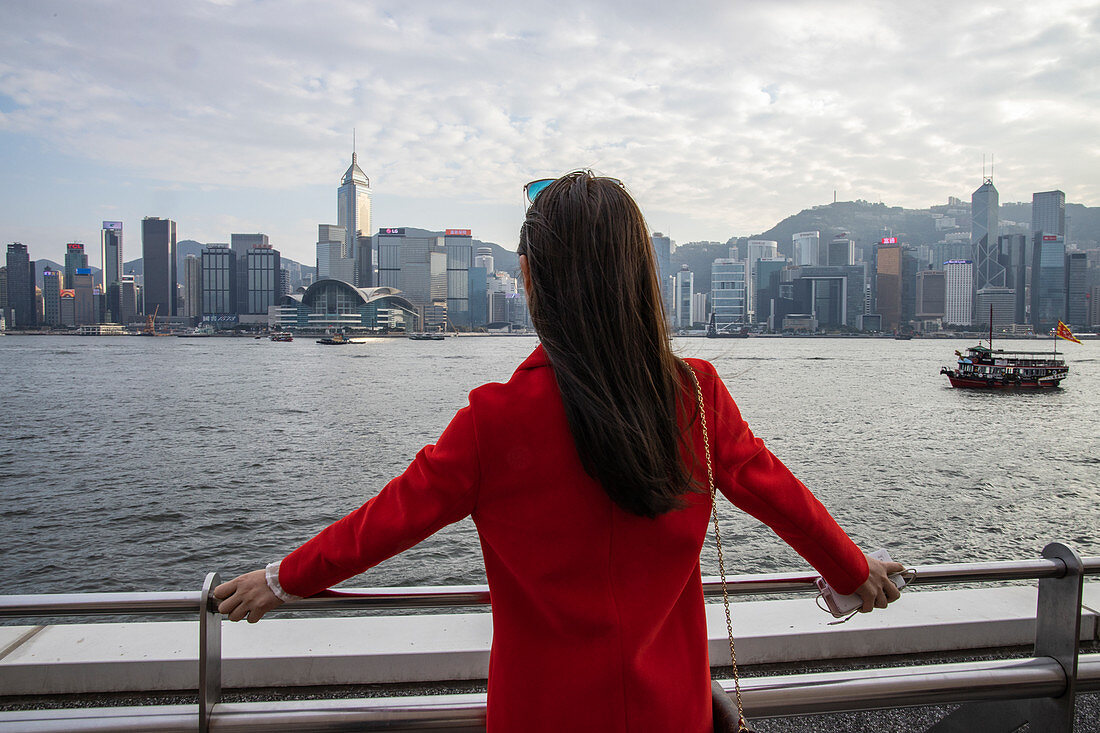 Frau mit rotem Blazer bewundert die Aussicht von der Tsim Sha Tsui-Seite des Victoria Harbour mit der Skyline von Hongkong in der Ferne, Hongkong, Hongkong, China, Asien