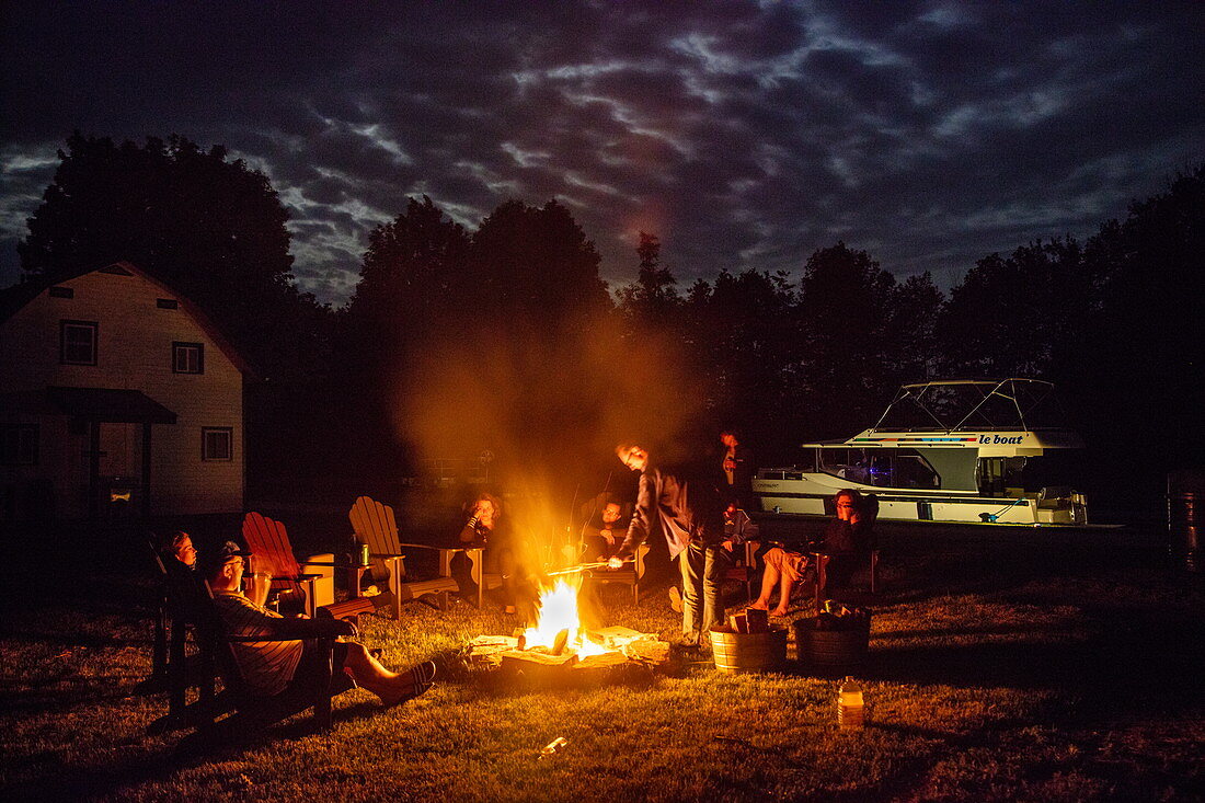 Menschen entspannen sich am Lagerfeuer bei Vollmond auf dem Parks Canada Campingplatz an den Schleusen Beveridge Locks am Fluss Tay River mit einem angedockten Le Boat Horizon Hausboot bei Nacht, nahe Lower Rideau Lake, Ontario, Kanada, Nordamerika
