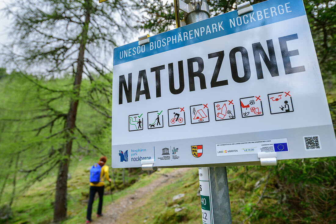 Sign nature zone with woman hiking out of focus in the background, Nockberge, Nockberge-Trail, UNESCO Nockberge Biosphere Park, Gurktal Alps, Carinthia, Austria