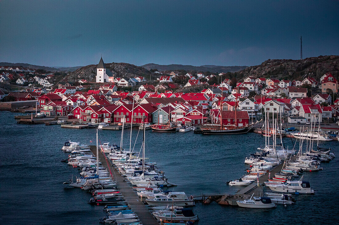 Rote Häuser mit Kirche und Booten im Hafen im Dorf Skärhamn auf der Schäreninsel Tjörn an der Westküste von Schweden, am Abend\n
