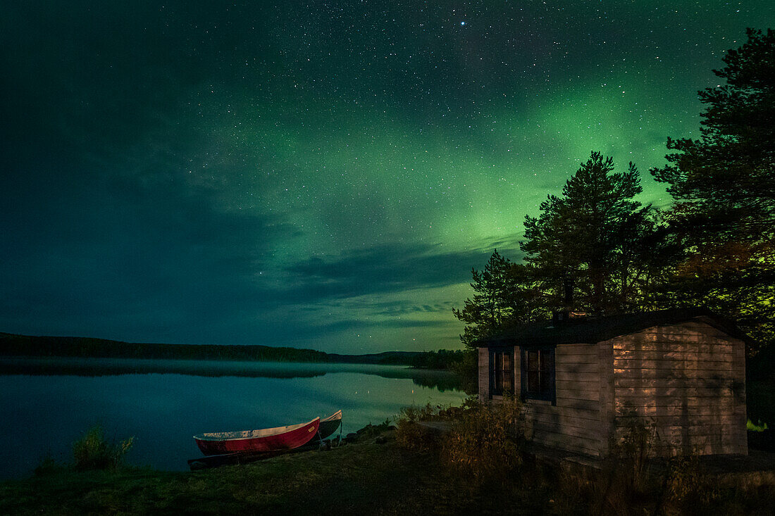 Polarlichter im Nachthimmel am Seeufer mit Hütte und Booten in Lappland, Schweden\n