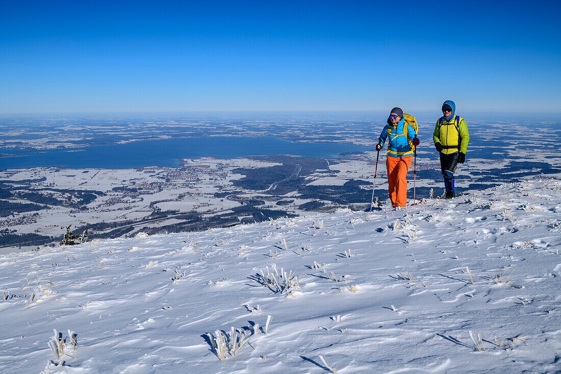 Man and woman hiking up over snow-covered slope to Hochgern, Chiemsee in the background, Hochgern, Chiemgau Alps, Upper Bavaria, Bavaria, Germany