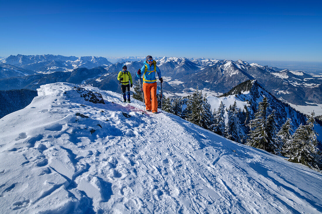 Mann und Frau beim Wandern steigen über verschneiten Hang zum Hochgern auf, Kaisergebirge und Chiemgauer Alpen im Hintergrund, Hochgern, Chiemgauer Alpen, Oberbayern, Bayern, Deutschland