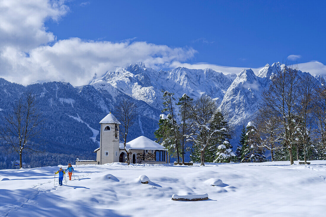 Man and woman hiking through snowy meadow with War Memorial Church and Wetterstein Mountains in the background, Kramerplateauweg, Garmisch, Ammergau Alps, Werdenfelser Land, Upper Bavaria, Bavaria, Germany