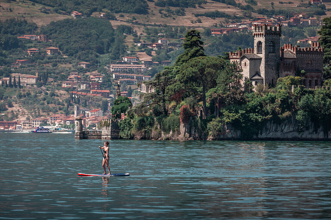 Woman paddles on SUP board in front of Castello della Isola di Loreto castle on island in Lake Iseo in Italy
