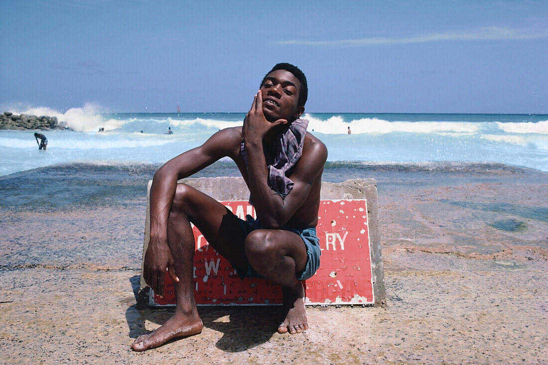 Junger Mann sitzt am Strand vor einem Warnschild, Barbados