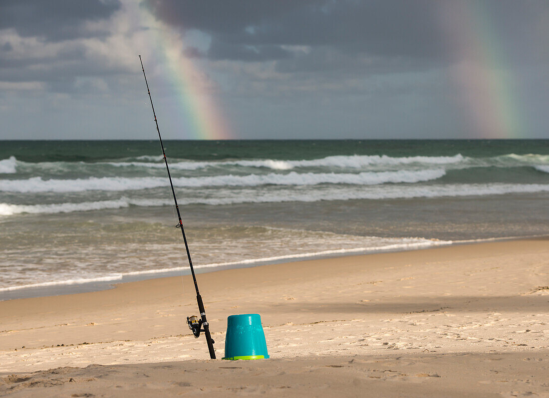 Surf Caster Angelrute im Sand und Regenbogen im stürmischen Himmel am Strand gesichert