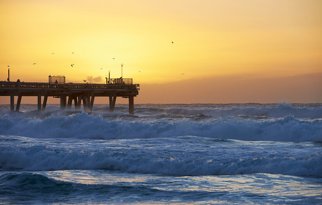 People walking and fishing on ocean pier as the sun set over the ocean