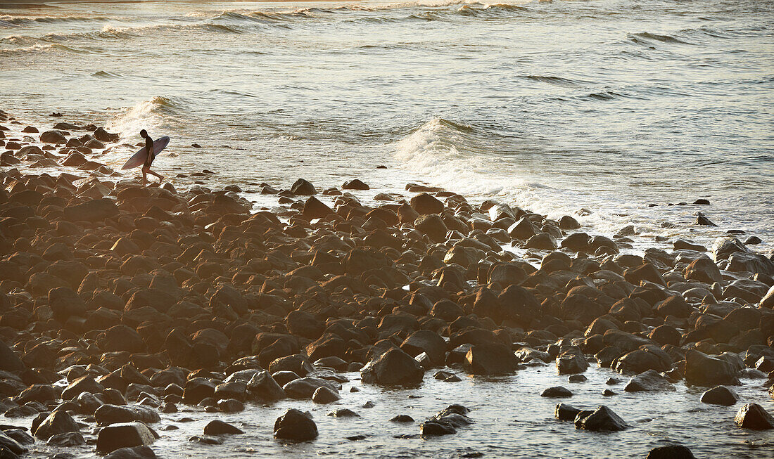 Lone Surfer carrying surfboard walking over rocks from the sea