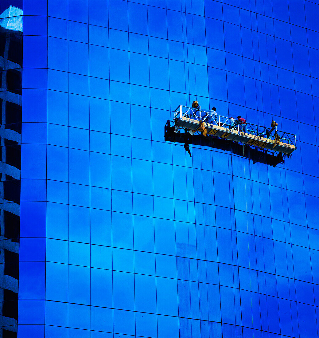 Two men in hanging scaffolding cleaning windows of high rise glass building with clouds reflecting in glass