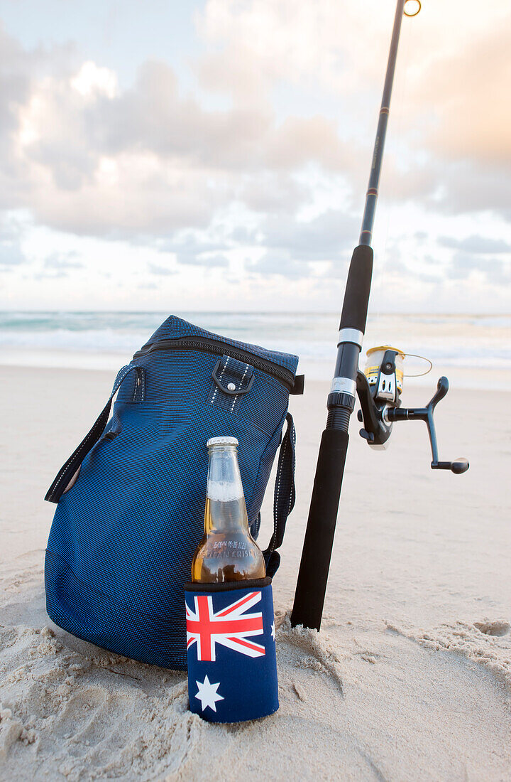 Bier im Stubbie-Halter mit australischer Flagge, Kühltasche und Angelrute am Strand am Australia Day
