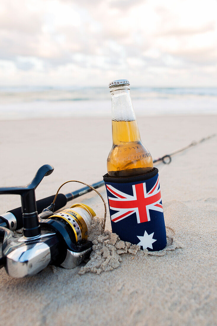 Beer in stubbie holder with Australian Flag next to Fishing rod on the sand at the beach