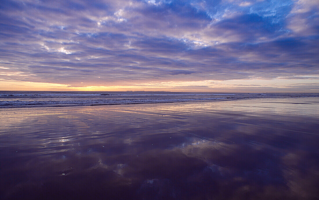 Wellen, die bei Sonnenaufgang auf nassen Sand rollen, reflektieren bunte Wolken am Himmel