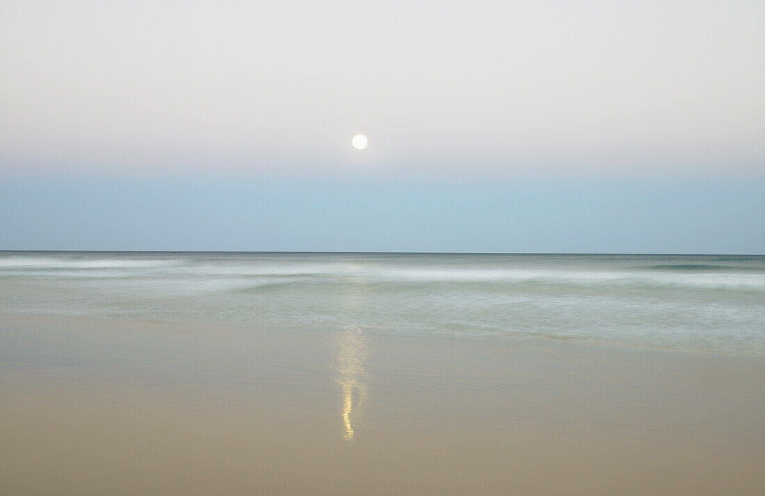 Moon rising in the evening sky over the ocean and reflecting in the sand