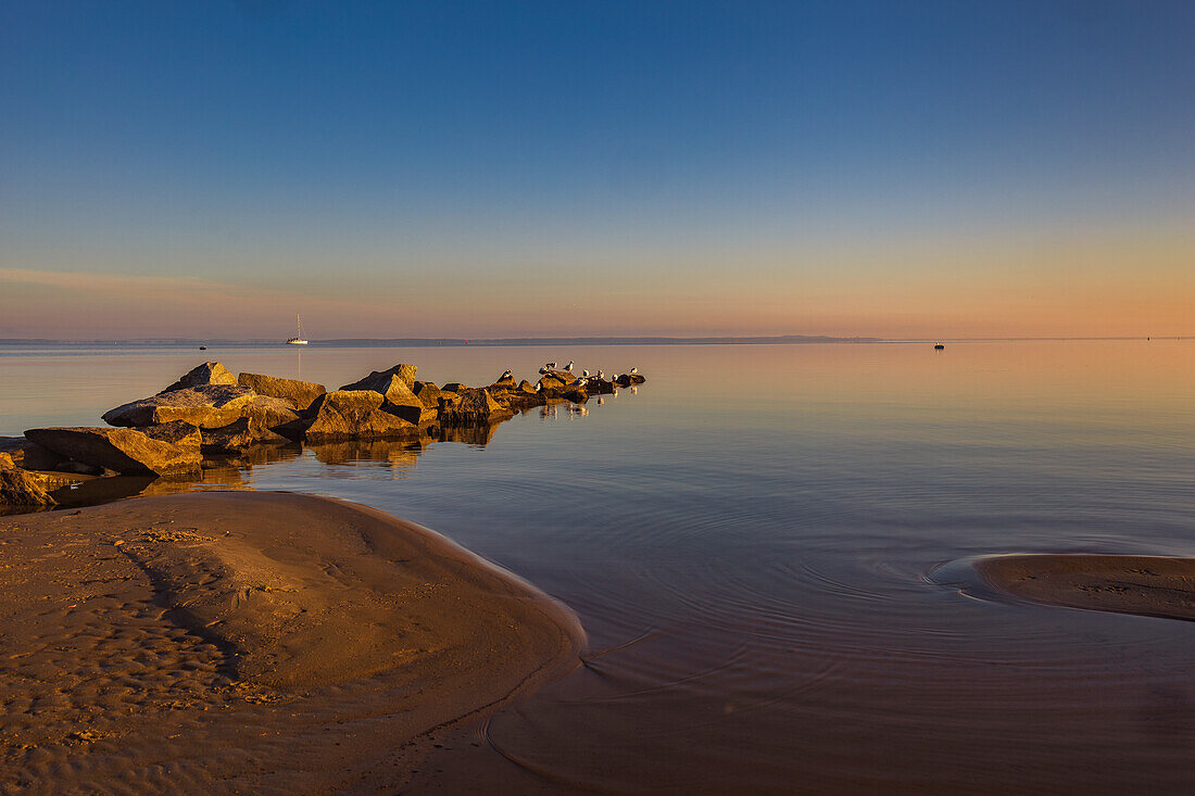 Ostseeküste mit Wellenbrecher bei Sonnenaufgang, im Hintergrund sieht man ein Segelboot. Stettiner Haff, Ostsee, Mecklenburg-Vorpommern, Deutschland, Europa