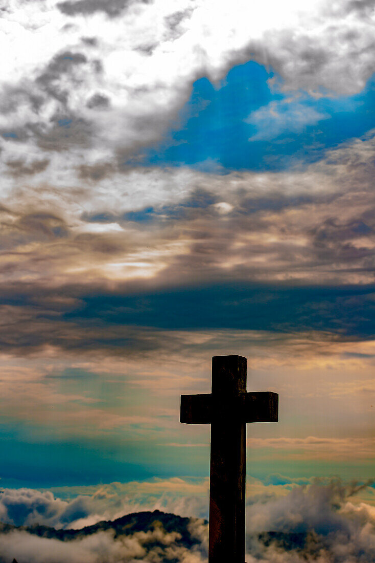 Religiöses Kreuz mit Blick auf die Berge und mit Wolkengebilde mit Sonnenlicht im Tessin, Schweiz