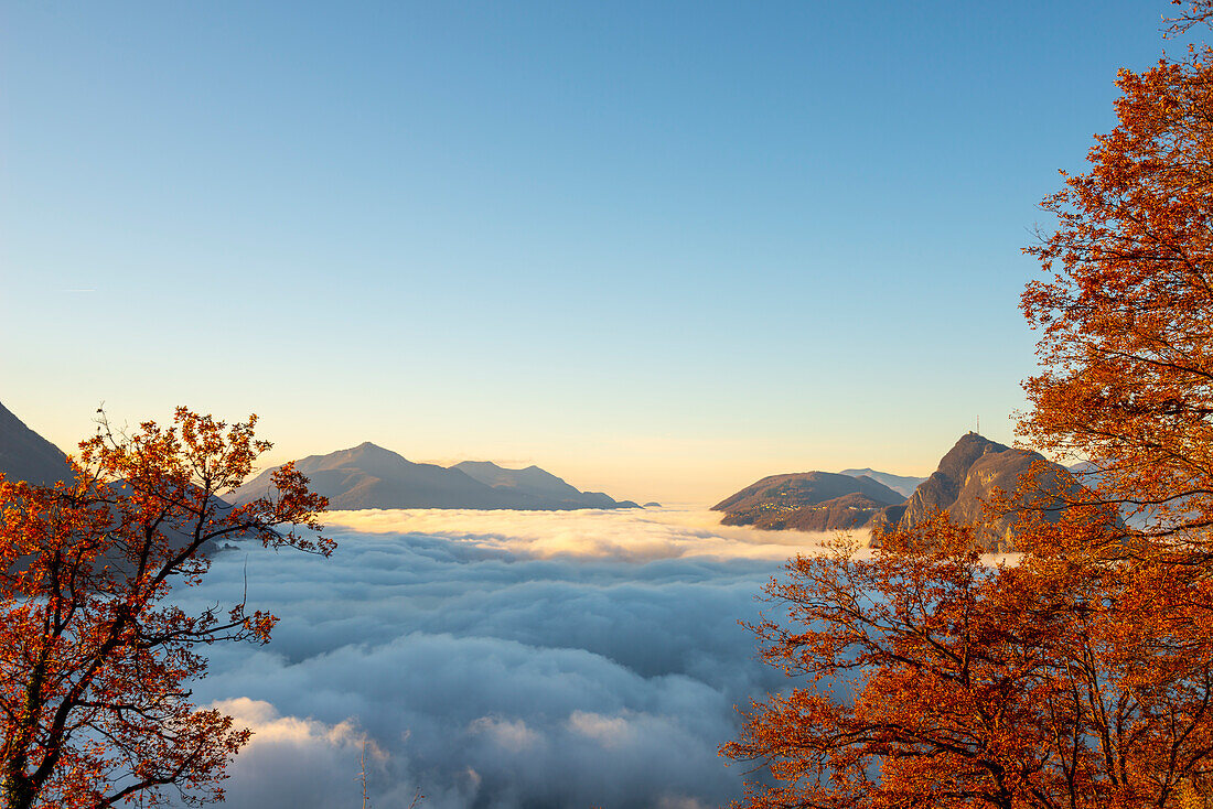 Berggipfel San Salvatore über Wolkengebilde mit einem Herbstbaum und Sonnenlicht mit klarem Himmel in Lugano, Tessin in der Schweiz