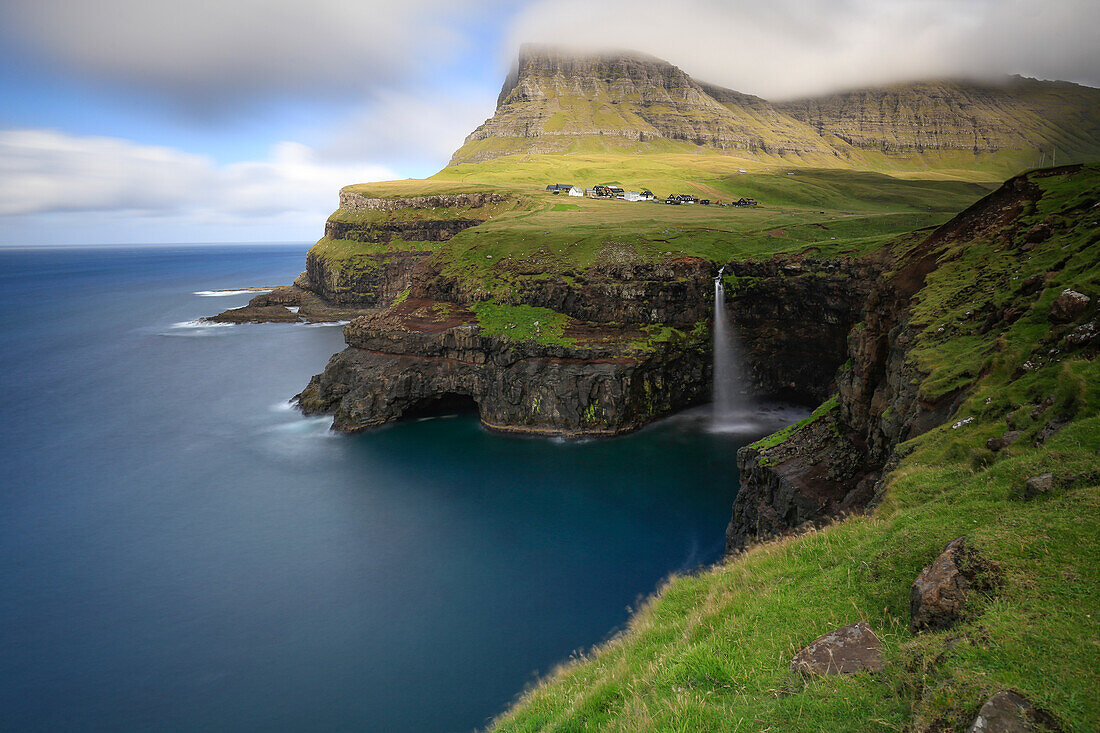 Blick auf Wasserfall Mulafossur und Ortschaft Gasadaluran an Steilküste von Vagar, Färöer Inseln. Wolken und blauer Himmel.