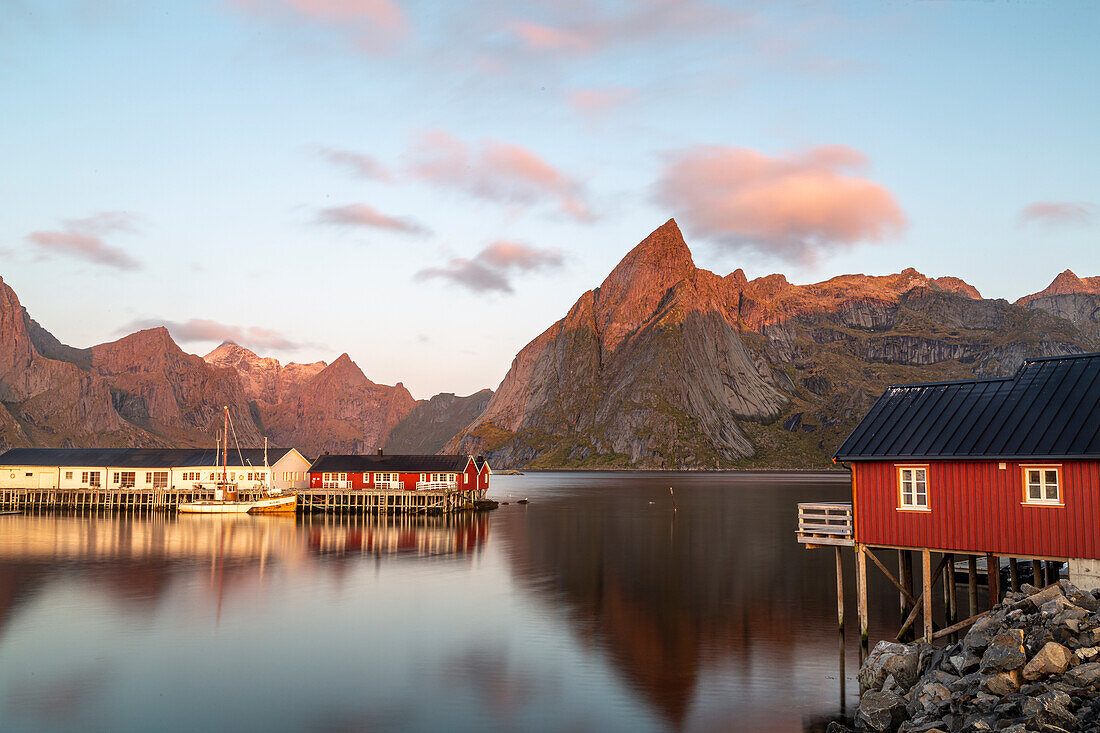 Fischerboote, rote Häuser im Hafen von Hamnoy, Moskenesoy, Lofoten, Norwegen. Sonnenlicht auf Bergpitzen.