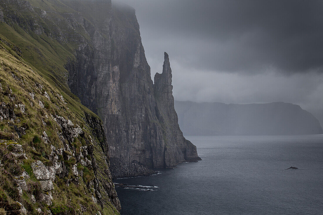 Blick entlang Steiküste auf Witches Finger, Felsnadel. Vagar, Färöer Inseln. Dunkle Regenwolken.