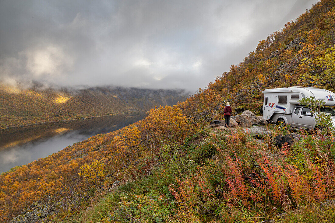 Person standing in front of camper van in autumn landscape, looking at Gryllefjord, Senja, Norway.