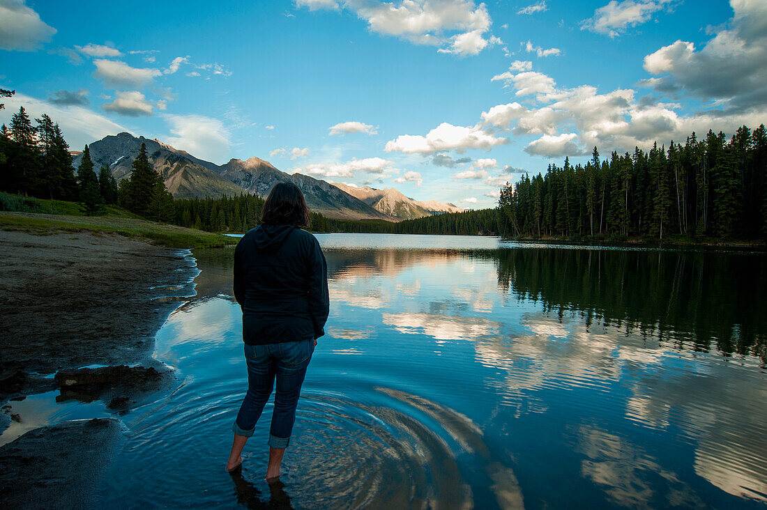 Frau, die im seichten Wasser des Johnson Lake im Banff National Park steht und Berge und ländliche Szene betrachtet, Kanada
