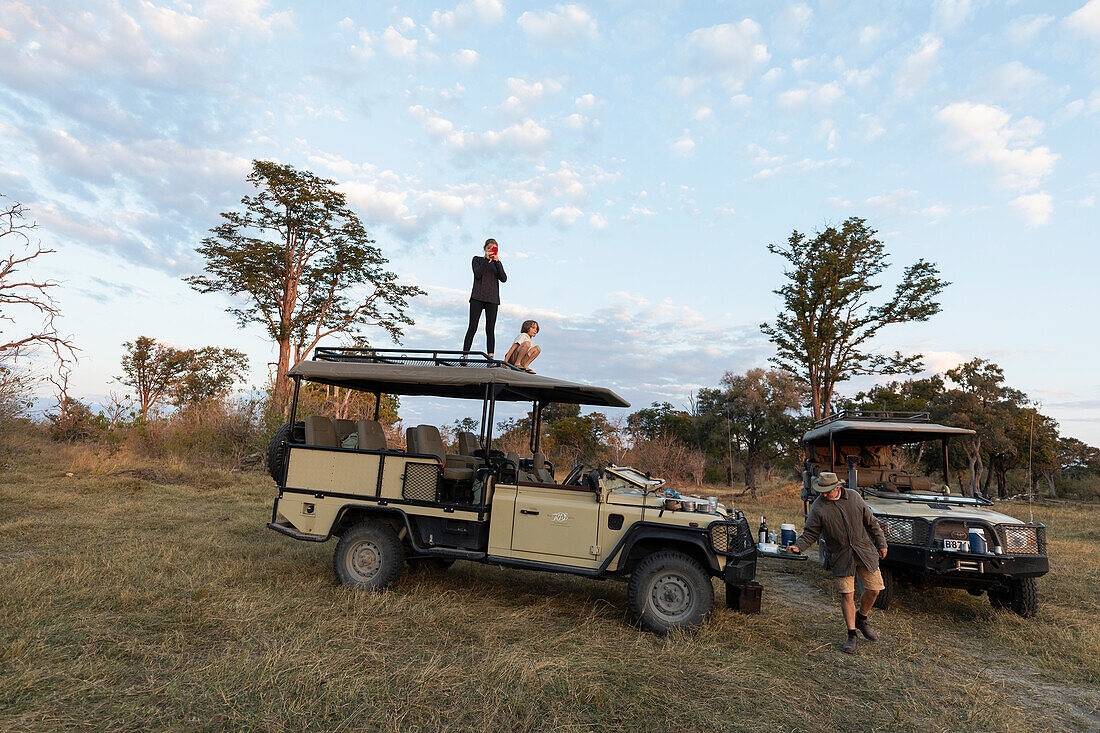 Zwei Kinder auf dem Dach eines Safarifahrzeugs bei einer Pirschfahrt bei Sonnenaufgang, Okavango-Delta, Botswana