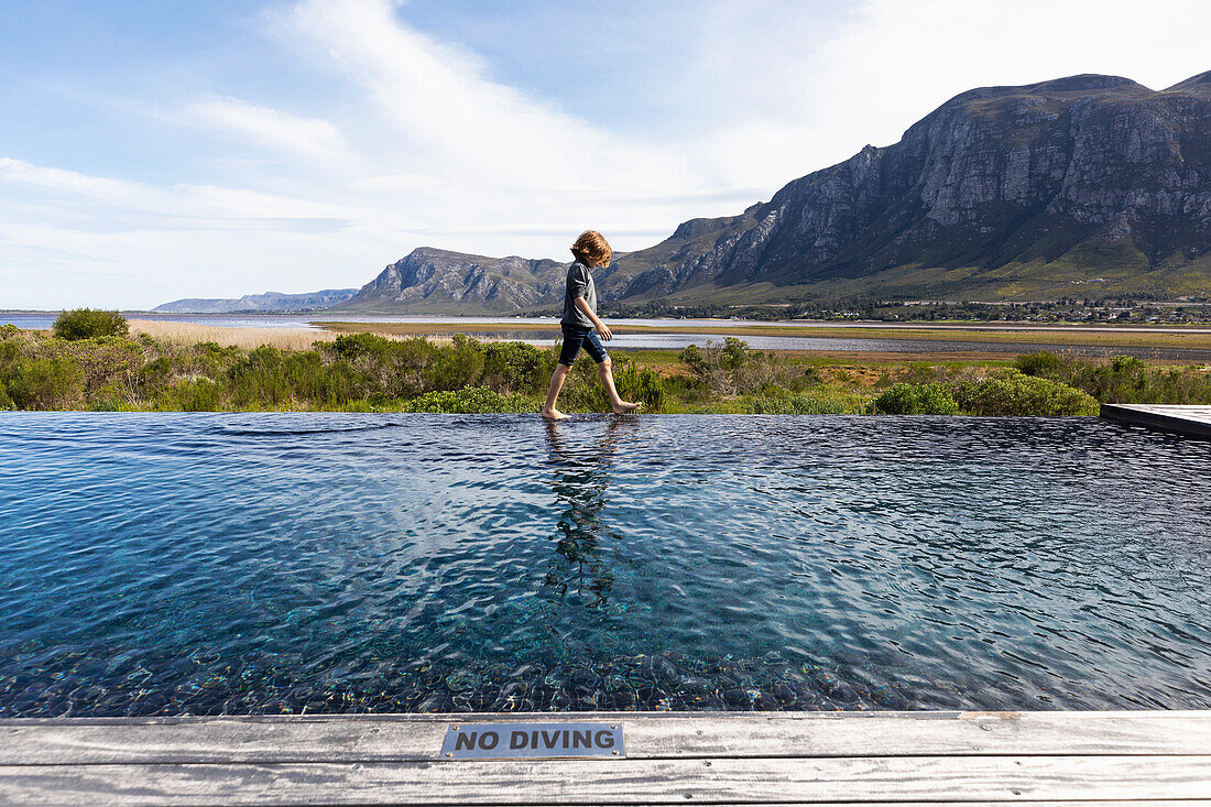Eight year old boy walking around the edge of an infinity pool, a mountain backdrop, Klein Mountains,South Africa