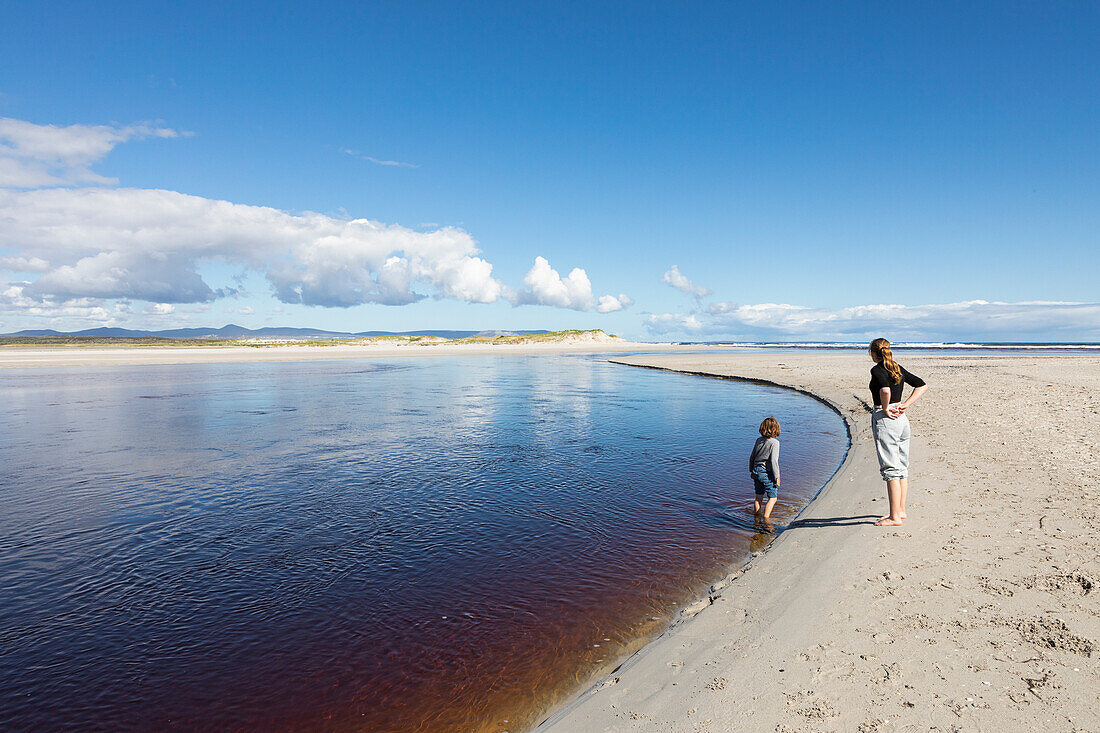 Teenage girl and a young boy at the edge of a wide water channel, on a sandy beach, Grotto Beach, South Africa