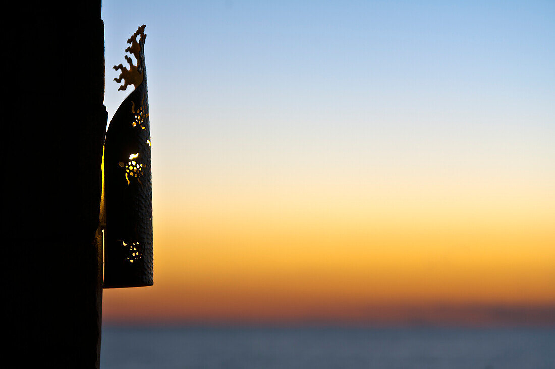 Ein Laternengitter an einer Wand, Silhouette gegen den Sonnenuntergangshimmel.