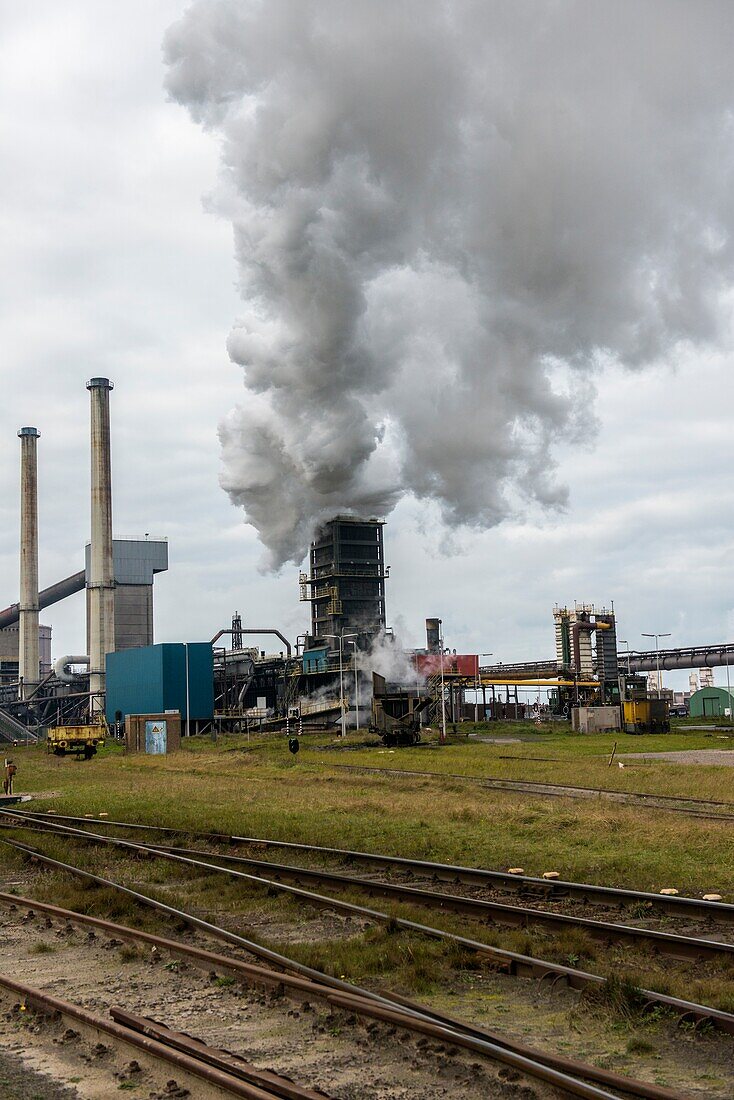 IJmuiden, Niederlande. Riesige, schwere Stahlproduktionsanlage und Industriegelände, in der verschiedene Arten von Stahl in einer CO2-emittierenden Fabrik hergestellt werden. Das Stahlwerk heißt Hoogovens und gehört Tata Steel, einem internationalen Konzern, der ebenfalls unter Umsatzeinbußen und immer höheren Anforderungen an die Unternehmensverantwortung in Bezug auf Arbeit und Klima leidet.