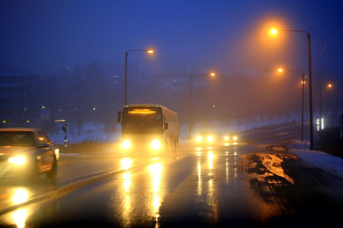 Scheinwerfer von Autos und einem Reisebus spiegeln sich an einem nebligen, blauen Winterabend in einem Vorort auf nasser Straße wider. Salo, Finnland.