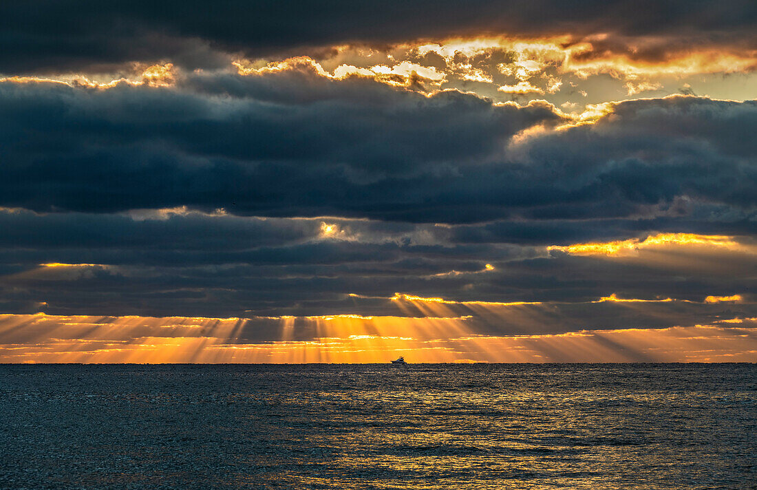 Sonnenstrahlen und goldene Wolken über dem Ozean bei Sonnenaufgang mit kleinem Fischerboot in der Ferne