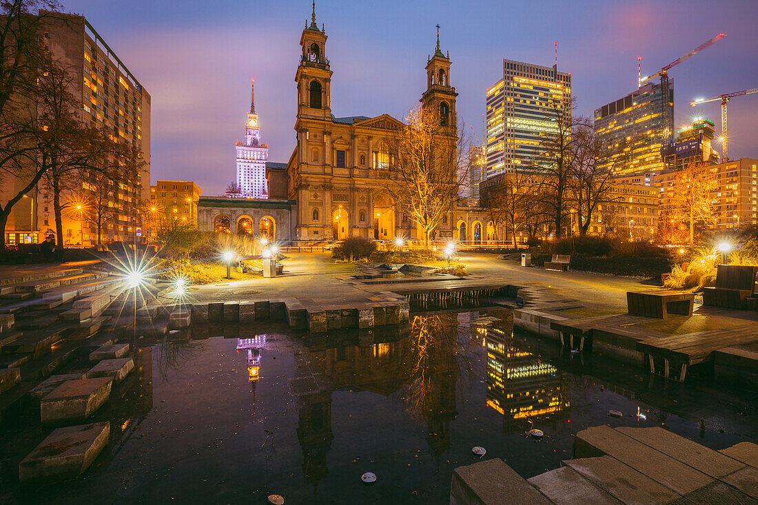 Polen, Masowien, Warschau, Kirche und beleuchtete Wolkenkratzer der Stadt, die sich nachts im Teich spiegeln