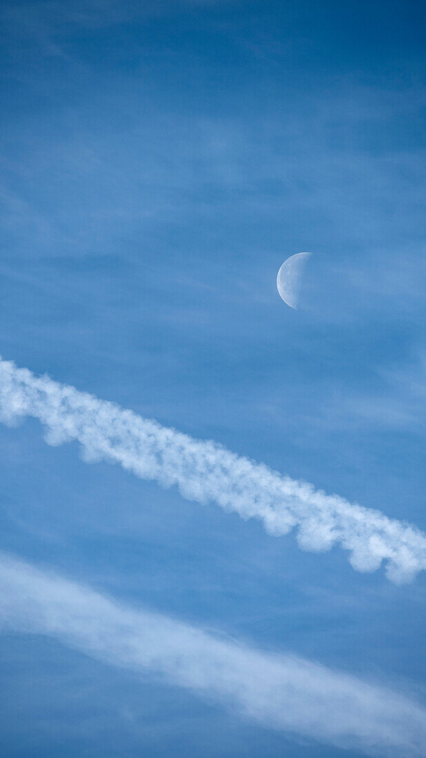 Flugzeugkondensstreifen und abnehmender Mond gegen blauen Himmel
