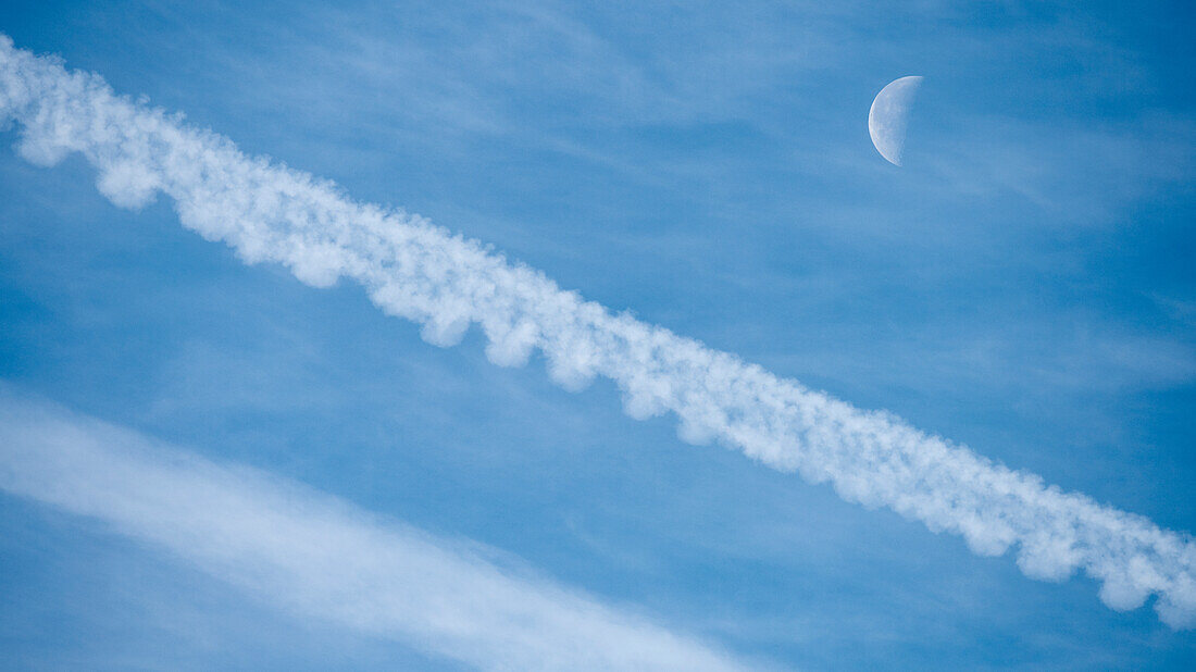 Flugzeugkondensstreifen und abnehmender Mond gegen blauen Himmel