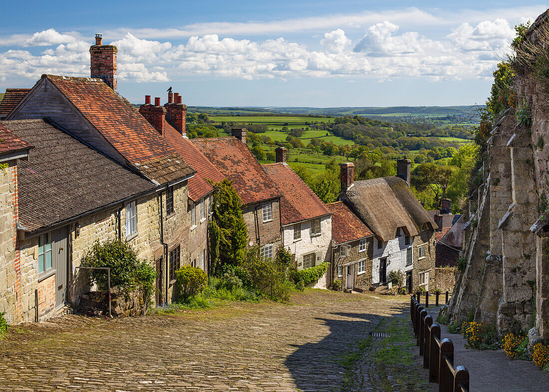 Gold Hill, gepflasterte Gasse, gesäumt von Cottages und Blick über die Landschaft, Shaftesbury, Dorset, England, Vereinigtes Königreich, Europa